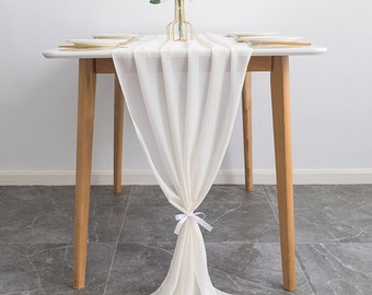 Könnte beinhalten: Ein weißer Chiffon-Tischläufer mit einer weißen Schleife in der Mitte. Der Läufer ist über einen Holztisch mit einer Vase mit rosa Blumen in der Mitte gelegt.