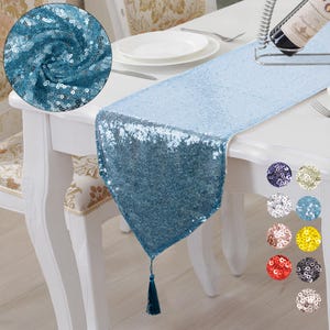May include: A light blue sequin table runner with a tassel. The runner is on a white table with a white tablecloth. The runner is 35.5 cm wide and 183 cm long. The runner is made of sequins and has a tassel on the end.