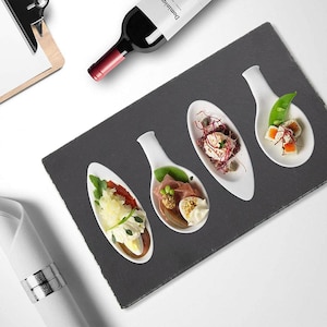 May include: Four white ceramic spoons with different food items on a black slate serving tray. The spoons are filled with a variety of toppings, including salad, ham, and cheese.