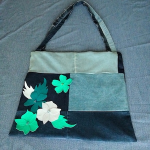 Peut inclure: Un sac cabas en jean bleu avec un design patchwork orné de fleurs en tissu vert et blanc. Le sac a une longue bandoulière et est fabriqué à partir de matériaux recyclés.