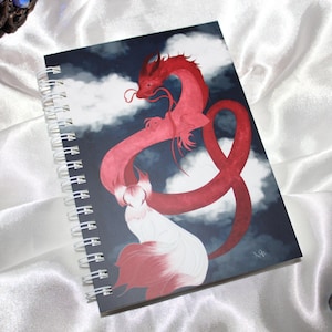 Peut inclure: Un carnet à spirales présentant une illustration numérique d'un long dragon rouge sinueux volant dans un ciel sombre et nuageux au-dessus d'une créature blanche à plusieurs queues avec des accents roses sur fond bleu marine.