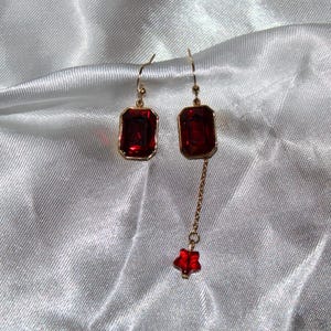 Op de afbeelding: Een paar gouden oorbellen met rode edelsteen druppels. De oorbellen hebben een rechthoekige rode edelsteen met een gouden zetting en een hangende stervormige rode edelsteen.