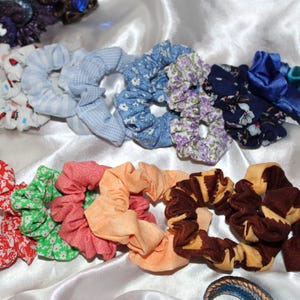 Peut inclure: Une collection de chouchous en tissu colorés avec différents motifs, notamment floraux, à carreaux et à pois. Les chouchous sont disposés en rangée sur un fond en tissu blanc.
