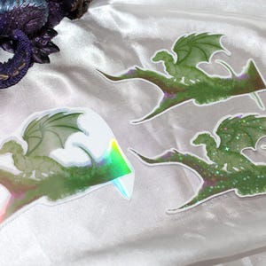 Peut inclure: Trois autocollants de dragon avec des ailes et des corps verts, perchés sur une base verte et marron. Un autocollant a un effet holographique. Une figurine de dragon violet est en arrière-plan.