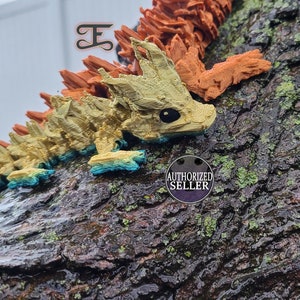May include: A small, colorful, 3D printed dragon figurine with a spiky, textured body. The dragon is painted in shades of orange, yellow, and blue. The dragon is perched on a tree trunk with green moss.