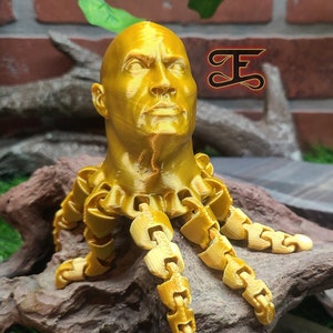 May include: A gold-colored 3D printed bust of a person with a chain-like design around the neck and extending out as arms. The bust is sitting on a brown wooden surface.