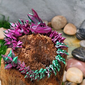May include: A pink and green iridescent crystal dragon sculpture on a wooden log surrounded by stones.