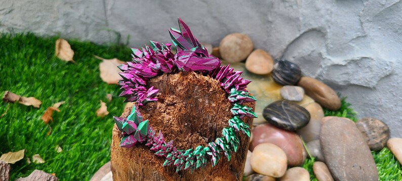 May include: A pink and green iridescent crystal dragon sculpture on a wooden log surrounded by stones.