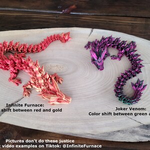 May include: Two origami dragons made of paper, one red and gold, the other green and purple. The dragons are on a wooden surface. The text "Infinite Furnace: Color shift between red and gold" is below the red dragon. The text "Joker Venom: Color shift between green and purple" is below the green dragon. The text "Pictures don't do these justice Check out video examples on Tiktok: @InfiniteFurnace" is at the bottom of the image.