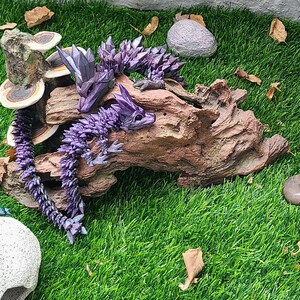May include: Three origami dragons made of paper, in shades of purple, green, and blue, are displayed on a wooden log and a tree stump. The dragons are surrounded by small stones and dried leaves.