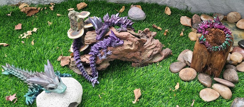 May include: Three origami dragons made of paper, in shades of purple, green, and blue, are displayed on a wooden log and a tree stump. The dragons are surrounded by small stones and dried leaves.