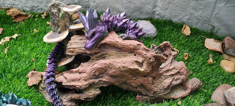 May include: A purple and silver crystal dragon figurine resting on a piece of driftwood with a mushroom growing on a nearby rock.