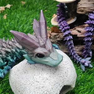 May include: A small, teal and purple dragon figurine with a spiky back and a white, textured egg-shaped base. The dragon has a light blue belly and a white, textured egg-shaped base.