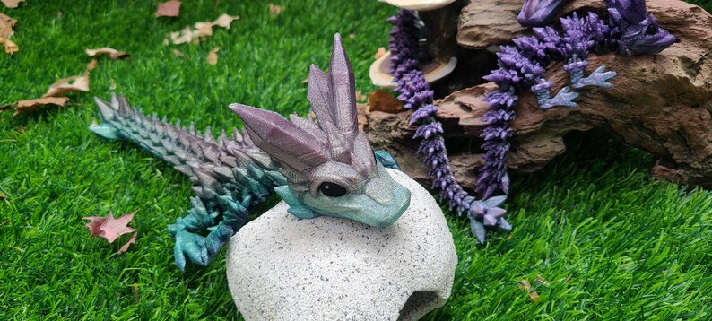 May include: A small, teal and purple dragon figurine with a spiky back and a white, textured egg-shaped base. The dragon has a light blue belly and a white, textured egg-shaped base.
