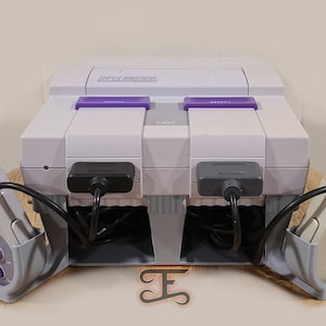 May include: A white Super Nintendo Entertainment System console with two gray controllers, each with a black cord. The console is sitting on a white stand with two gray controller holders. The console has a purple power button and a gray cartridge slot.