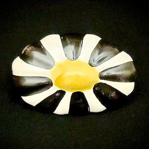 May include: A decorative ceramic dish with a daisy-like design. The dish features alternating black and white petals surrounding a yellow center. The dish is set against a black background.