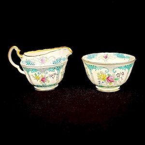 May include: A cream and turquoise porcelain creamer and sugar bowl set. The creamer has a curved handle and spout, while the bowl is round. Both pieces feature a floral design with gold trim. The set is against a black background.