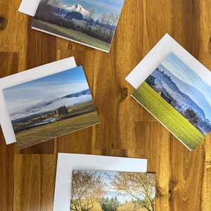 May include: Four white envelopes with different landscape photographs inside. The photos include a mountain range, a foggy lake, a field of trees, and a field of flowers.