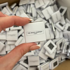 May include: A small, white square product with the text "THE WHITE COMPANY LONDON" printed in black. The product is held in a hand, with many similar items visible in the background.