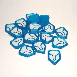 Könnte beinhalten: Ein Haufen blau-weißer Spielsteine. Jeder Spielstein hat einen blauen Umriss und ein weißes Innenteil mit einem stilisierten Symbol. Die Spielsteine liegen auf einer weißen Oberfläche verstreut.