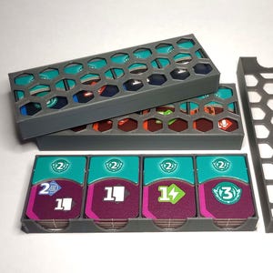 Puede incluir: Un juego de cuatro porta tarjetas de plástico gris con un diseño de panal. Los porta tarjetas están diseñados para guardar y organizar las tarjetas de un juego de mesa. Las tarjetas son visibles a través del plástico transparente.