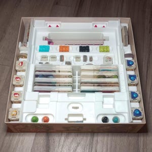 May include: A board game organizer with various components, including cards, tokens, and game pieces. The organizer is made of white plastic and cardboard, with compartments for each game element. The game pieces are in various colors, including green, orange, and blue.