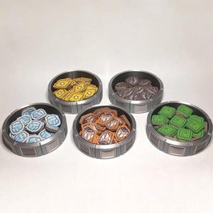 May include: Four round containers with lids, each filled with small, colorful tokens. The containers are made of a silver-colored metal and have a textured design. The tokens are in shades of blue, yellow, brown, and green.