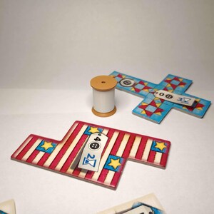 May include: A wooden cotton reel and several game pieces. The game pieces are in the shape of a cross and a flag, with red and white stripes, blue stars, and tags with numbers and symbols.