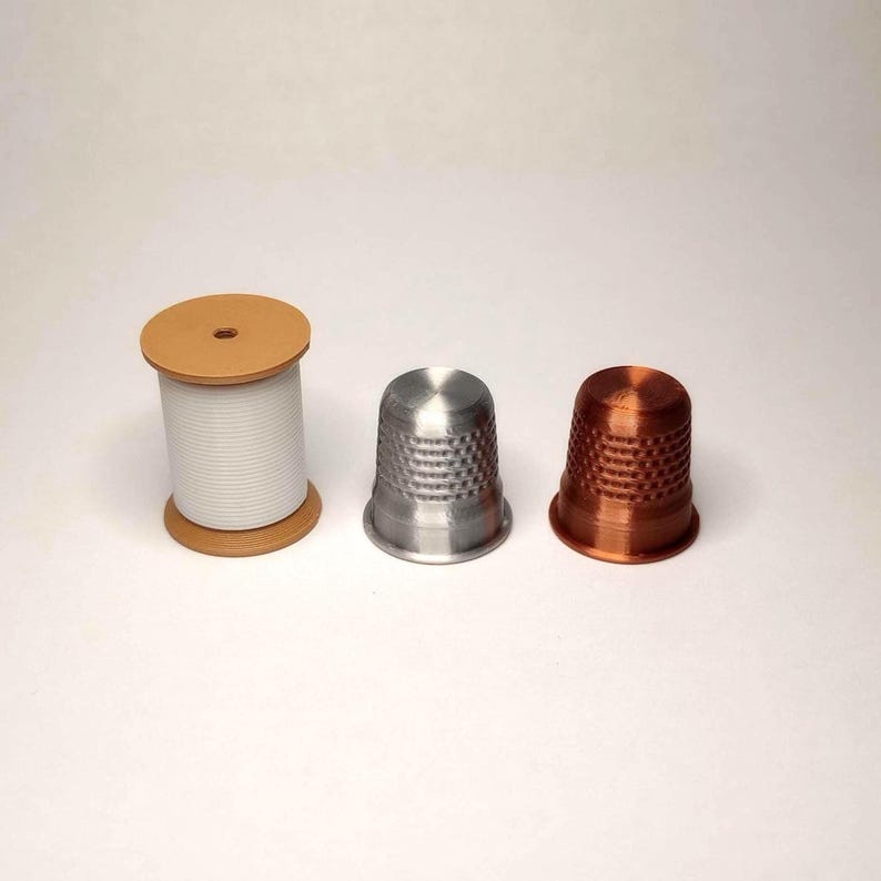 May include: Three sewing tools on a white surface. A spool of white thread with brown ends stands to the left. In the center is a silver thimble, and to the right is a copper-colored thimble. The thimbles have a dimpled texture.