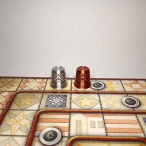 May include: A silver and a copper thimble are placed on a board game. The game board features a patterned design with geometric shapes and a brown border. Small, circular game pieces are also present.