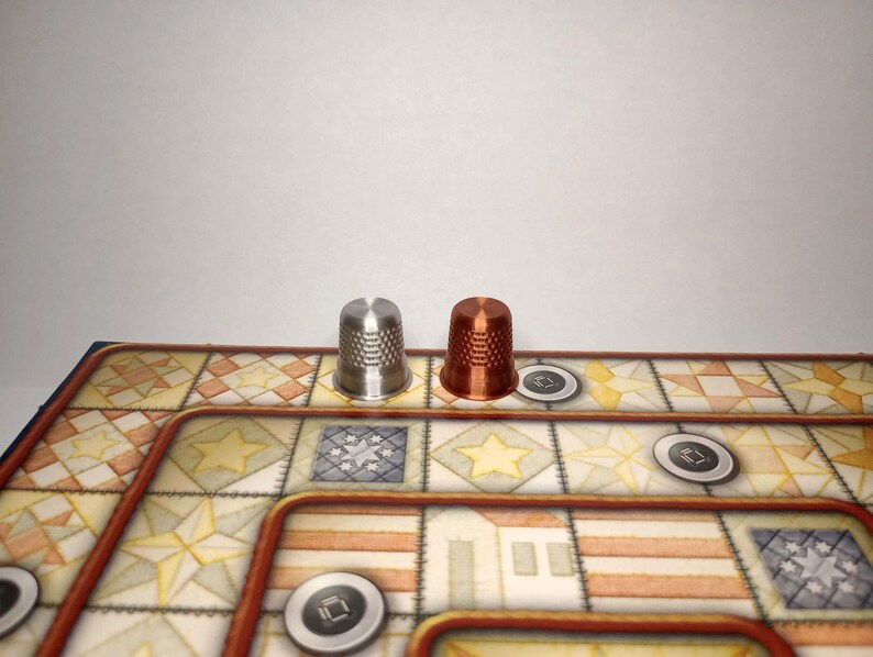 May include: A silver and a copper thimble are placed on a board game. The game board features a patterned design with geometric shapes and a brown border. Small, circular game pieces are also present.