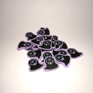 May include: A pile of small, black ghost-shaped charms with a white swirl design on a light purple background. These charms are likely for crafting or jewelry making, perfect for Halloween-themed projects.