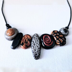 May include: A handcrafted necklace with a selection of stone-effect beads in black, brown, grey, and tan. The beads display distinct textures and patterns, threaded on a black cord.