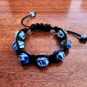 May include: A handmade bracelet featuring blue and white marbled beads strung on a black cord. The bracelet has an adjustable sliding knot closure. The beads are approximately 0.5 inches in diameter. The bracelet is displayed on a wooden surface.