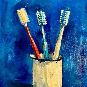 May include: An oil painting on canvas depicting three toothbrushes standing upright in a cylindrical white cup against a deep blue background. The toothbrushes have red, blue, and white handles. The artist's name, Ana Doolin, is signed at the bottom.