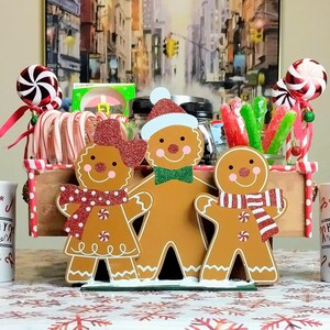 Puede incluir: Una cesta de regalo con temática de casa de jengibre con tres figuras de jengibre, bastones de caramelo y dos tazas que dicen "Holiday Candy Homemade".