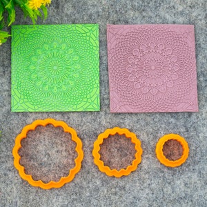 Puede incluir: Dos alfombrillas de estampado en silicona verde y morado con intrincados diseños de mandala. Tres cortadores de galletas de plástico naranja con bordes festoneados.