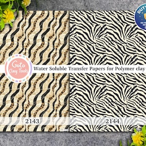 May include: Two sheets of water soluble transfer paper for polymer clay. The left sheet features a beige and black wavy animal print pattern labeled 2143. The right sheet displays a black and white zebra stripe pattern labeled 2144. Text reads Goto Clay Tools.