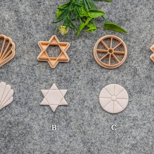 May include: Eight cookie cutters in various shapes, including a seashell, a Star of David, a wheel, and a square. The cutters are made of a light brown plastic and are arranged in two rows of four. The cutters are on a grey textured surface.