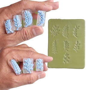 Puede incluir: Un conjunto de herramientas para nail art. La imagen muestra varios sellos de uñas rectangulares transparentes con diseños de hojas azul claro. También se ve un molde rectangular verde con varios patrones de hojas. Las herramientas están diseñadas para crear nail art botánico.