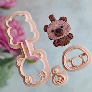 May include: A selection of light pink bear-shaped cookie cutters and a brown bear-shaped fondant decoration. The cutters include a bear head, a bear body, and a steering wheel shape. The fondant bear has stitched details.