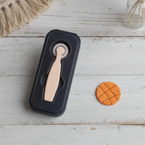 May include: A black and beige rotary cutter with a beige handle and a circular blade. A small, round, orange cookie with a crosshatch pattern is next to the cutter. The cutter is in a black case.