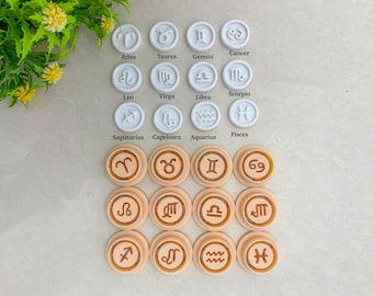 May include: A set of 12 white and beige round zodiac sign cookie cutters. The white cutters are arranged in two rows of six, with the zodiac signs Aries, Taurus, Gemini, Cancer, Leo, Virgo, Libra, Scorpio, Sagittarius, Capricorn, Aquarius, and Pisces. The beige cutters are arranged in three rows of four, with the same zodiac signs.