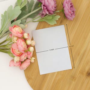 May include: A rectangular white card measuring 4 inches by 5.5 inches, positioned next to a bouquet of pink and cream-colored artificial ranunculus flowers with green stems and leaves, all arranged on a light brown wooden surface.