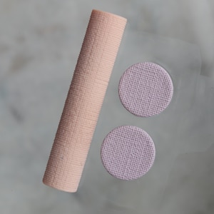 Puede incluir: Un objeto cilíndrico texturizado de color melocotón claro se coloca junto a dos objetos circulares texturizados de color lavanda. Los artículos están sobre una superficie translúcida, lo que sugiere un producto cosmético o de belleza.