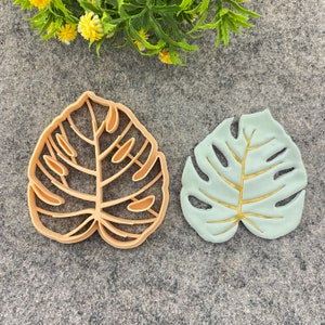 Puede incluir: Dos cortadores de galletas con forma de hojas de monstera. Uno es un cortador de plástico marrón claro y el otro es un cortador de plástico azul claro con un diseño de hoja dorada.