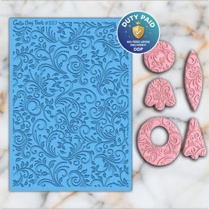 Puede incluir: Un molde de silicona azul Goto Clay Tools #557 con un detallado diseño floral. También se muestran varias formas de arcilla rosa con el mismo diseño floral, incluyendo un círculo, una lágrima, una campana, un anillo y un triángulo.