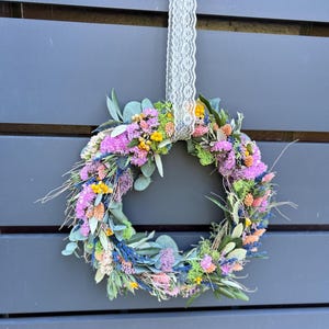 May include: A vibrant floral wreath composed of dried flowers in pink, yellow, blue, and orange hues. The wreath is hung by a white lace ribbon against a grey backdrop. A decorative item for the home.