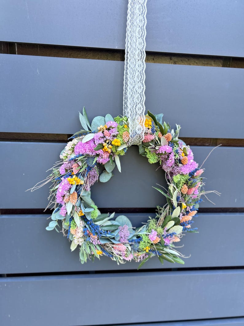 May include: A vibrant floral wreath composed of dried flowers in pink, yellow, blue, and orange hues. The wreath is hung by a white lace ribbon against a grey backdrop. A decorative item for the home.