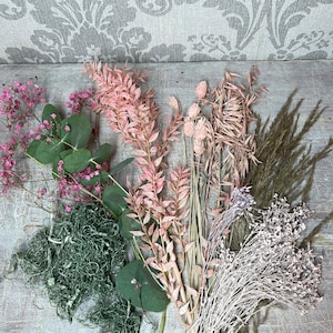 May include: An assortment of dried flowers and greenery, including pink baby's breath, eucalyptus, and feathery grasses. The arrangement features various textures and shades of pink, green, and brown, perfect for crafting or home decor.